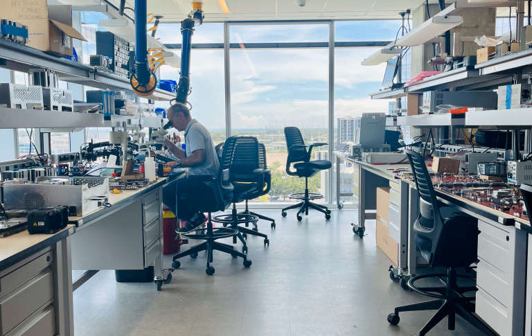 Annapurna Labs: A man looks through a microscope in a cluttered lab space, chairs are haphazardly set through the room, with counter space a premium, a polished off-white floor leads to large windows, which display the Austin skyline that is blue filled with clouds.